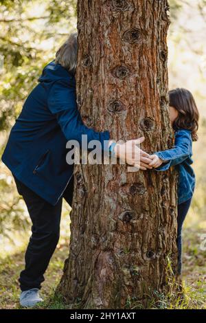 Vue latérale de la petite fille pleine longueur en veste en denim et homme âgé avec des cheveux gris debout dans la forêt autour du bois et tenant les mains sur le dos flou Banque D'Images