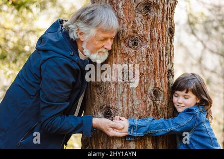 Vue latérale d'une jolie petite fille en veste denim et d'un homme âgé aux cheveux gris debout dans la forêt autour du bois et tenant les mains sur un arrière-plan flou Banque D'Images