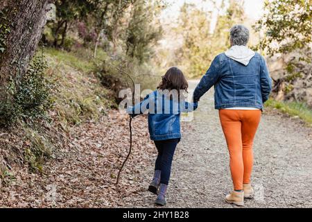 Corps complet de grand-mère anonyme tenant la main avec petite-fille avec bâton marchant sur le chemin couvert de feuilles sèches dans la forêt Banque D'Images