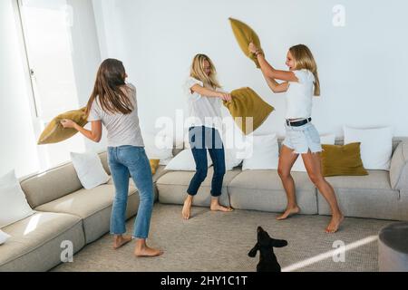 Un corps complet d'amis heureux pieds nus se battant avec des oreillers tout en s'amusant ensemble dans un salon léger à la maison Banque D'Images
