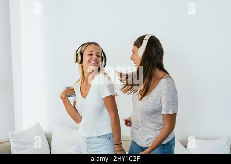 Des amies positives écoutent de la musique dans un casque sans fil tout en dansant et en s'amusant dans un salon léger à la maison Banque D'Images