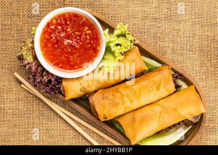 Vue de dessus de délicieux petits pains de printemps frits faits maison dans un plateau en bois avec une sauce douce au piment rouge dans une tasse en céramique blanche et des légumes verts. Banque D'Images