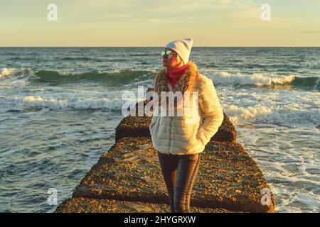Une femme aux cheveux blonds dans un chapeau et un manteau de fourrure marche le long de la mer le long d'un brise-lames en pierre Banque D'Images