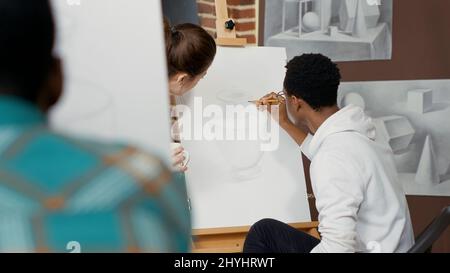 Un jeune enseignant parle avec l'étudiant de la conception de dessin dans un atelier de création, en utilisant des outils artistiques pour créer des œuvres d'art sur toile avec chevalet. Pratique de classe d'art pour dessiner un croquis de vase pour la croissance. Banque D'Images