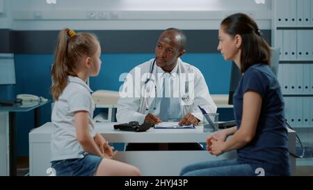 Médecin parlant aux patients et prenant des notes sur l'ordinateur pour préparer le document de contrôle avec le médicament de prescription. Medic ayant la conversation avec la petite fille et l'adulte dans le cabinet médical. Banque D'Images