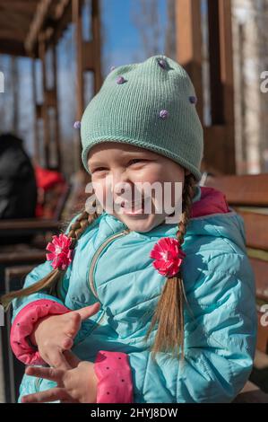 Portrait d'un enfant positif assis sur un banc de parc. Fille en train de rire avec de longues queues de porc et des arcs en forme de fleur. Un enfant de cinq ans content. Ressort Banque D'Images