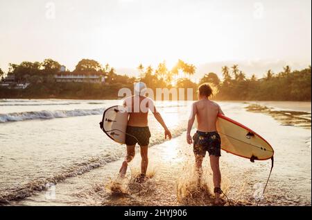 Père avec un fils adolescent marchant avec des planches de surf près de la plage de sable de l'océan avec des palmiers sur fond éclairé par le soleil de coucher de soleil. Ils aiment l'été Banque D'Images