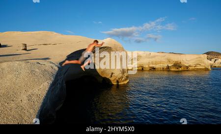 Lumière du soir, l'homme saute des rochers dans l'eau, roche tuff bizarre, ciel bleu, nuage gris-blanc, bleu mer foncé, Baie de Sarakiniko, île de Milos, Cyclades Banque D'Images