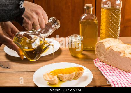 Détail des mains versant de l'huile d'olive extra vierge sur une tranche de pain sur une assiette blanche sur une table rustique en bois. Banque D'Images