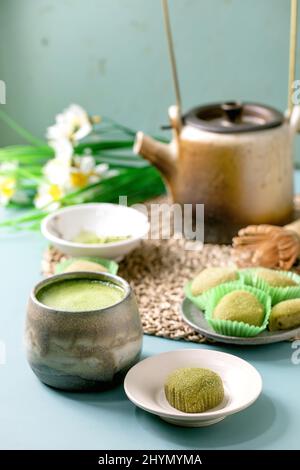Dessert de riz traditionnel asiatique mochi de matcha vert doux avec une tasse de thé matcha moussé sur une table bleue. La vieille théière et les jonquilles printanières fleurissent au dos Banque D'Images