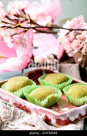 Gros plan du dessert de riz traditionnel asiatique mochi de matcha vert doux dans un plat de céramique rose sur une table bleue. Fleurs roses de printemps à l'arrière-plan. Banque D'Images