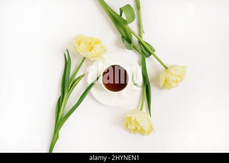 Une tasse en porcelaine blanche avec thé noir sur une soucoupe entourée de tulipes jaunes de printemps sur un fond blanc. Le concept du printemps, des femmes et de la mère Banque D'Images