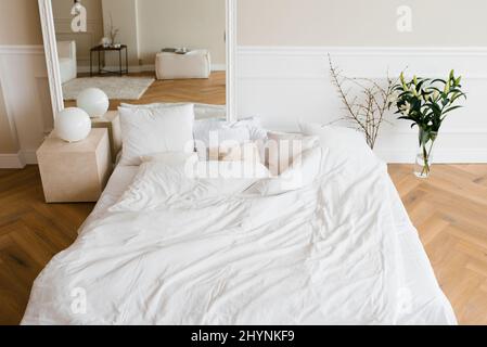 Un lit double avec draps blancs et oreillers beige dans une chambre scandinave lumineuse, un lit au sol et des fleurs dans un vase Banque D'Images