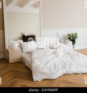 Un lit double avec draps blancs et oreillers beige dans une chambre scandinave lumineuse, un lit au sol et des fleurs dans un vase, un grand miroir dans un cadre Banque D'Images