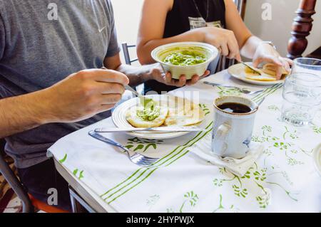 homme caucasien assis à la table à manger à côté de sa petite amie manger et remplir un arepa avec guacamole avec une tasse de café sur la table, piquant Banque D'Images