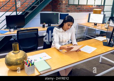 Jeune femme d'affaires biraciale lisant des documents tout en étant assise au bureau dans un bureau moderne Banque D'Images