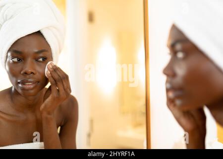 Reflet de la femme afro-américaine frottant l'éponge sur le visage dans la salle de bains à la maison Banque D'Images