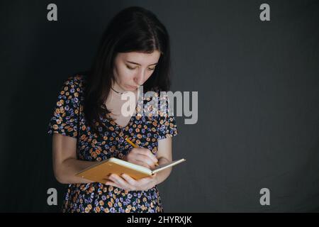 une jeune femme arménienne ukrainienne prend des notes avec un stylo dans son carnet sur fond gris Banque D'Images