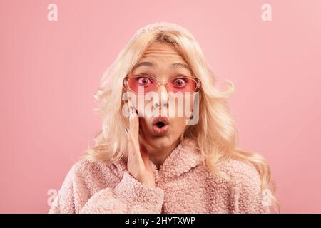 Offre OMG. Une jeune femme choquée avec la main sur la joue et regardant l'appareil photo dans l'excitation, fond rose, prise de vue en studio Banque D'Images