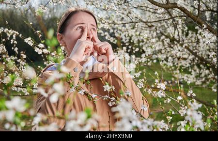 Femme allergique souffrant d'allergies saisonnières au printemps, posant dans un jardin fleuri au printemps. Jeune femme éternuant parmi les arbres en fleurs. Concept d'allergie printanière Banque D'Images