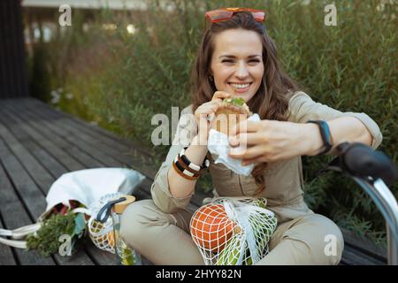 souriante femme de 40 ans dans l'ensemble avec sac fourre-tout, sac à ficelle, scooter et sandwich manger tout en étant assis à l'extérieur de la ville. Banque D'Images