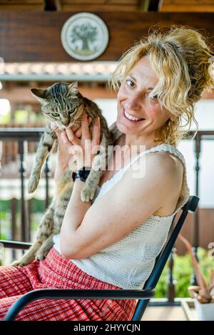 portrait d'une jeune femme blonde caucasienne mûre souriant avec son chat à la maison sur le porche assis dans une chaise à bascule. Concept de style de vie. Banque D'Images