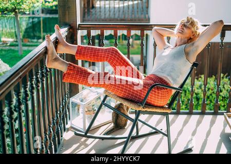 portrait d'une jeune femme blonde caucasienne mûre se détendant à la maison sur le porche assis dans une chaise à bascule. Concept de style de vie. Banque D'Images