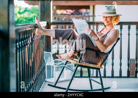 Portrait d'une jeune femme blonde de race blanche mature dans son 50s portant un chapeau et lisant un journal et assis dans une chaise à bascule dans une posture détendue Banque D'Images