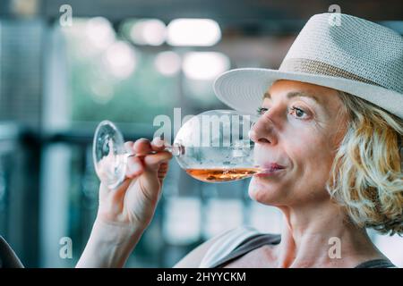 Portrait d'une jeune femme blonde de race blanche mature dans son 50s portant un chapeau et goûtant un verre de vin sur le porche d'une maison de campagne. Style de vie c Banque D'Images