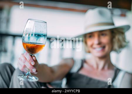 Portrait d'une jeune femme blonde de race blanche mature dans son 50s portant un chapeau et appréciant un verre de vin sur le porche d'une maison de campagne. Style de vie Banque D'Images