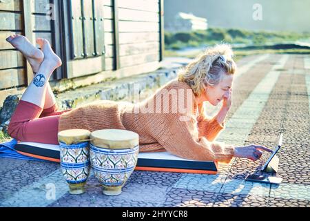 Jeune femme blanche mûre et blonde se détendant à l'extérieur avec une tablette par temps ensoleillé. Plage de Berria, Cantabrie, Espagne, Europe. Banque D'Images