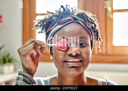 Gros plan Portrait amusant d'une jeune femme noire avec une coiffure d'afro plaisantant avec un lolipop intérieur à la maison, riant. Concept de style de vie. Banque D'Images