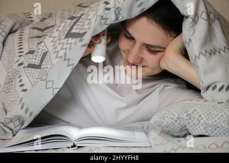 Une femme heureuse lit un livre couché sous une couverture la nuit et éclaire une lampe de poche sur les pages. Banque D'Images