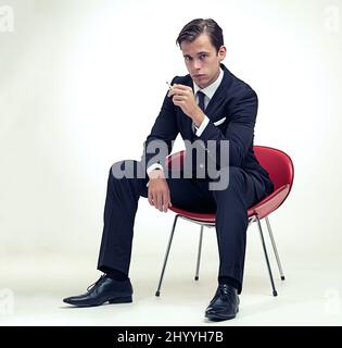 Suave fumeur. Un portrait en studio d'un beau jeune homme en costume à fines rayures assis sur une chaise. Banque D'Images