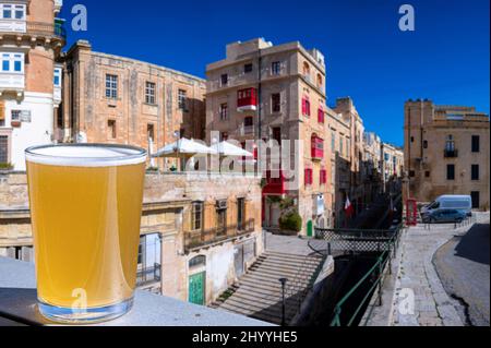 Verre de bière légère avec vue sur la passerelle et la rue étroite avec des escaliers à la Valette, Malte. Banque D'Images