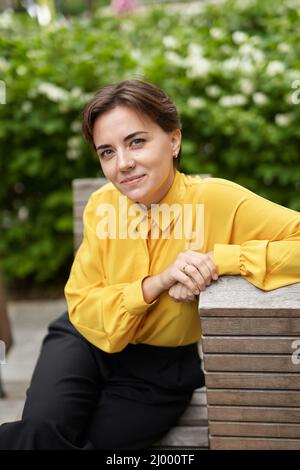 Portrait en gros plan d'une femme d'affaires attirante, d'un directeur ou d'un directeur immobilier assis sur un banc dans le parc en centre-ville, portant un chemisier jaune regardant directement dans l'appareil photo. Image de haute qualité Banque D'Images