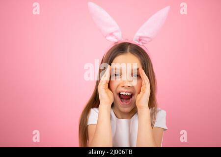 Gros plan d'une petite fille mignonne portant des oreilles de lapin sur la tête regardant l'appareil photo avec la bouche largement ouverte et couvrant son visage sur deux côtés avec Banque D'Images