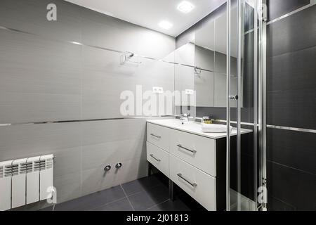 Toilettes avec armoire de salle de bains, évier en résine blanche, radiateur en aluminium, armoire avec miroir et cabine de douche avec carreaux sombres Banque D'Images