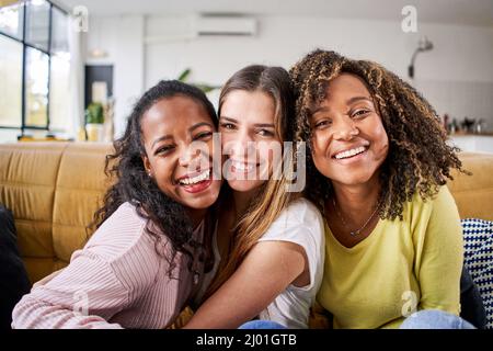 Trois courses mixtes des femmes heureuses embrassant sourire - femmes drôles ensemble célébrant assis sur le canapé du salon en regardant le selfie d'appareil photo. Banque D'Images