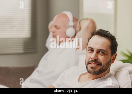 Homme assis souriant à la canera tandis que son père ou son partenaire s'assoit à côté de lui écoute de la musique sur des écouteurs Banque D'Images