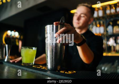 Vue en contre-angle du barman professionnel qui fait un cocktail alcoolique coloré debout derrière le comptoir du bar dans la boîte de nuit moderne sombre. Banque D'Images