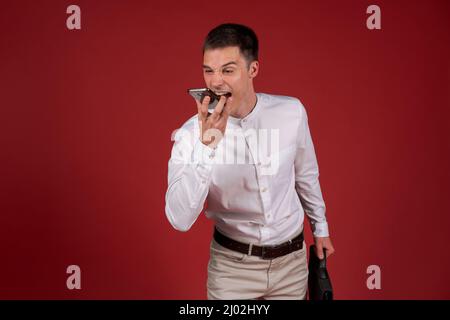 Un homme en colère dans une chemise blanche et un pantalon léger avec un sac à documents dans sa main hurle sur un téléphone portable sur le haut-parleur.Un homme stressé et déprimé sur fond rouge.Homme émotionnel Banque D'Images