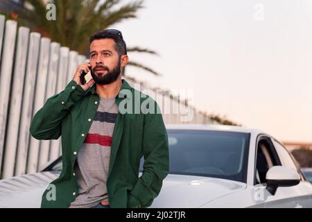 jeune homme barbu attrayant assis sur le capot de voiture parlant sur le téléphone mobile, concept de technologie de loisirs et de communication, espace de copie pour le texte Banque D'Images