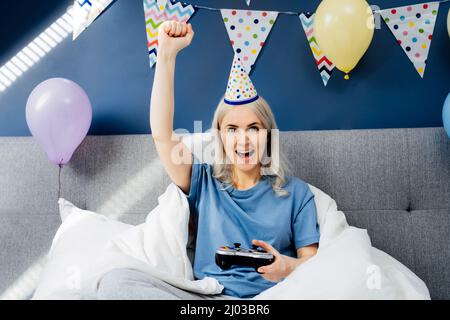 Femme émotive et excitée en chapeau d'anniversaire assis sur son lit confortable avec contrôleur et jouer à un jeu vidéo. Idées de fête d'anniversaire à la maison. Temps en ligne, vir Banque D'Images