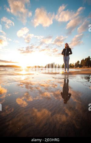 Un jeune athlète souriant de brunette marche pieds nus sur une plage de sable en jeans et un chemisier noir, en appréciant le coucher du soleil et les derniers rayons chauds. Sotkamo, Finla Banque D'Images