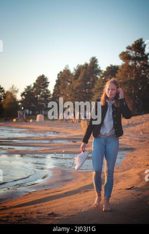 Un jeune athlète souriant de brunette marche pieds nus sur une plage de sable en jeans et un chemisier noir, en appréciant le coucher du soleil et les derniers rayons chauds. Sotkamo, Finla Banque D'Images