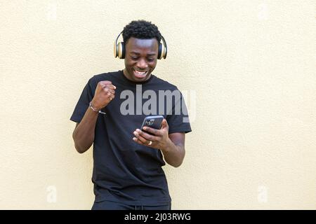 Jeune homme afro-américain enthousiaste, vêché de vêtements et de casques décontractés, se moque de sourire heureux tout en lisant un bon message debout Banque D'Images