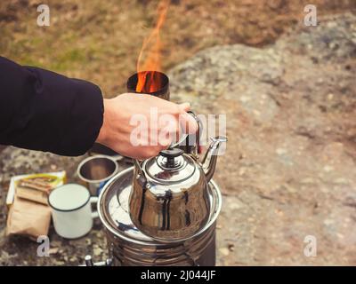 La main d'un homme tient la théière chromée sur le fond d'un feu de cheminée samovar. Thé boire dans la nature le jour de l'automne Banque D'Images