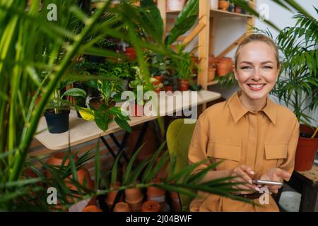 Portrait d'une fleuriste féminine gaie en ligne discutant avec le client en utilisant le téléphone mobile dans la boutique florale, regardant l'appareil photo. Banque D'Images