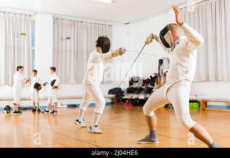 Jeunes tireurs regardant le duel d'escrime avec des feuilles de deux athlètes professionnels en salle de gym. Banque D'Images